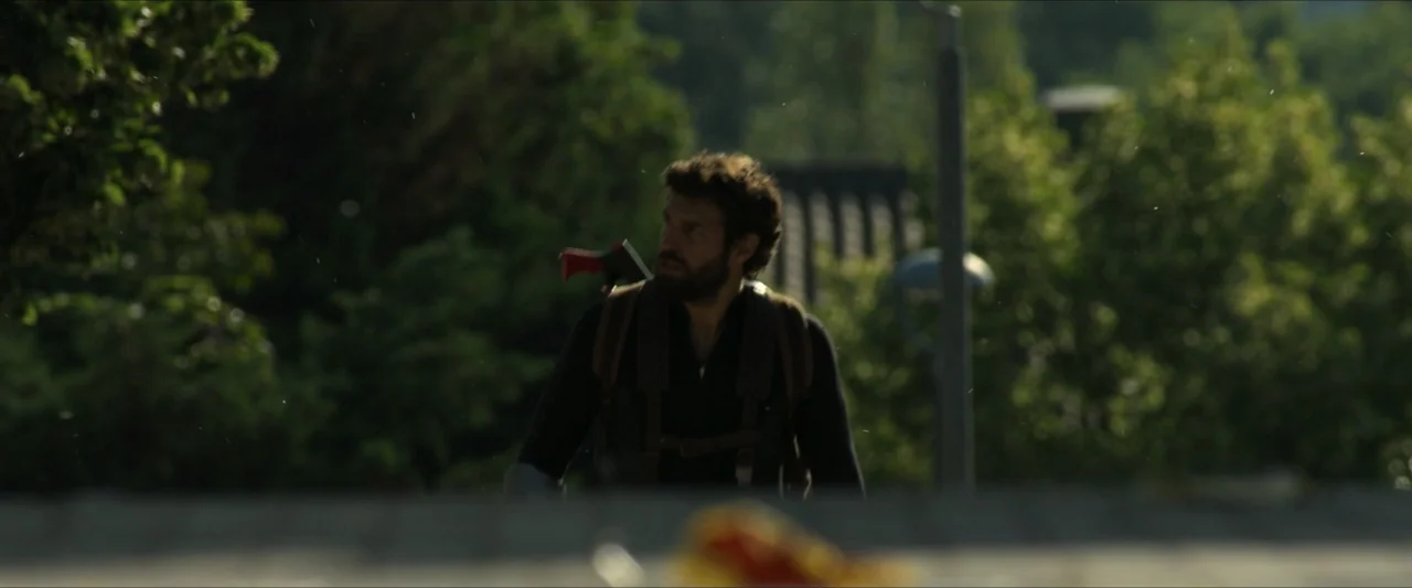 Apocalypse Z: The Beginning of the End (2024) movie still: medium — A sunny residential street with green trees and a blurred foreground object. A…