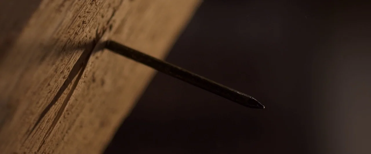 You’re Next (2013) movie still: closeup — A dark workshop space features a close view of a single iron nail. The nail is driven partially into a…