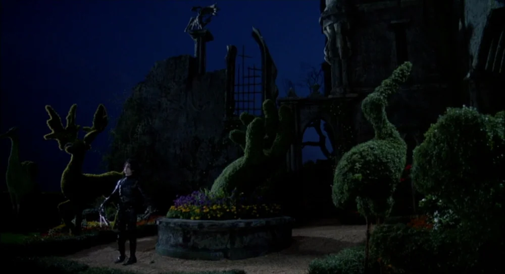 Edward Scissorhands (1990) movie still: wide — A dark night at a gothic mansion with large, elaborate topiary shapes. A thin man in a leather outfit…