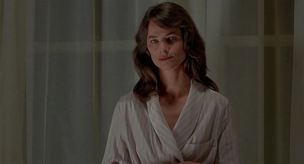 The Verdict (1982) movie still: medium — A woman stands indoors in front of large glass window panes covered by sheer curtains. She is framed from…