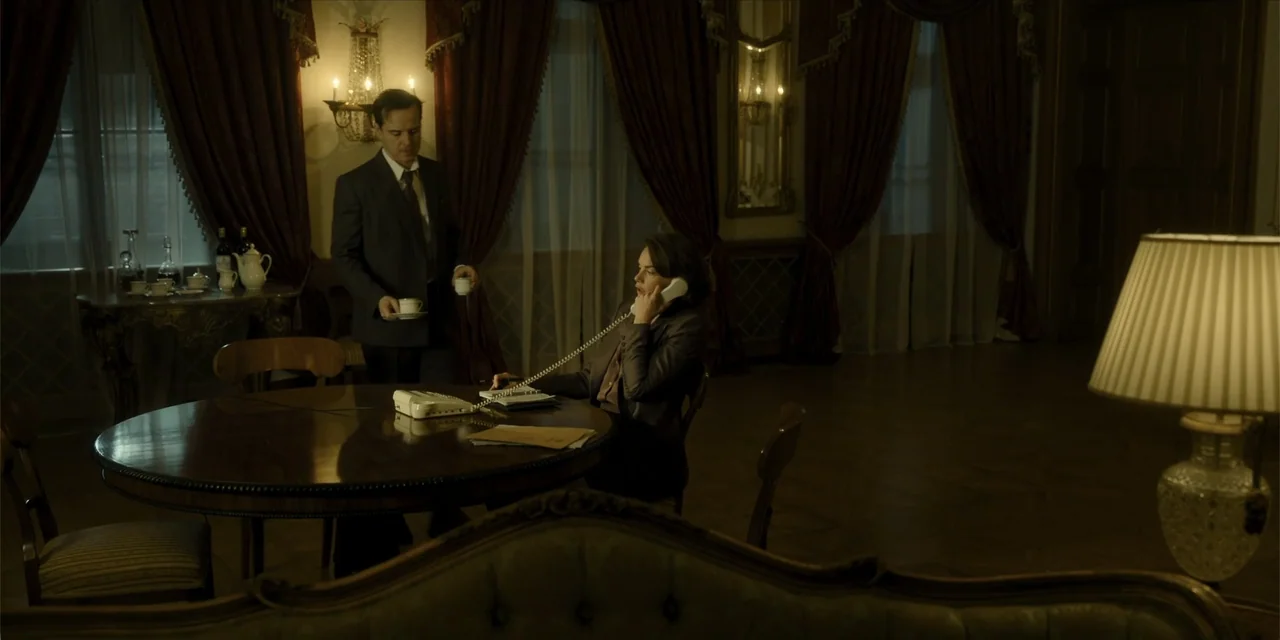 Oslo (2021) movie still: medium — A dimly lit hotel suite with dark wood furniture and floor-to-ceiling heavy curtains. A man serves tea to a woman…