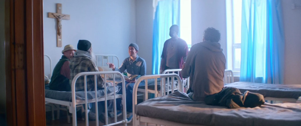 Room For Rent (2017) movie still: wide — A spare dormitory room features multiple white metal cots and a wooden crucifix on wall. Five men gather in…