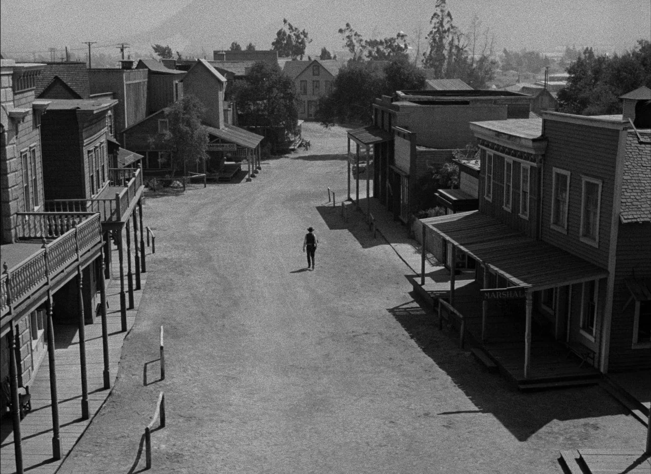 High Noon (1952) movie still: wide — A quiet, dusty town with wooden storefronts lining a wide, unpaved dirt path. A lone figure walks down the…