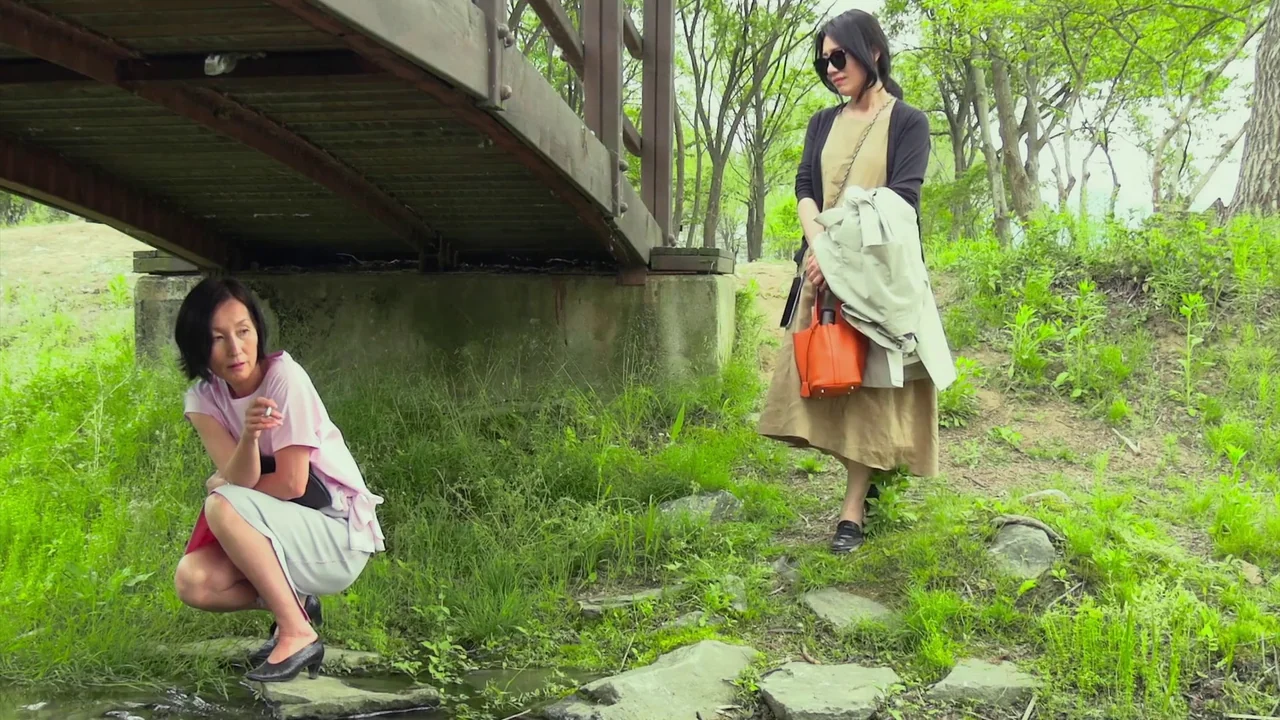In Front Of Your Face (2021) movie still: fullbody — A grassy area beneath a simple wooden bridge in a leafy park. One woman squats near the water's…