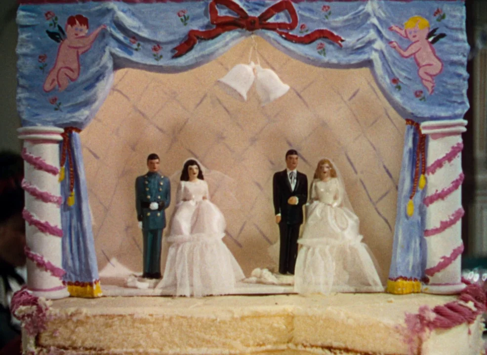 Calamity Jane (1953) movie still: closeup — A decorative cake topper features two miniature wedding couples under a pastel-painted fabric arch. The…