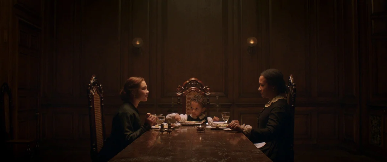 Lady Macbeth (2016) movie still: wide — A dimly lit room with dark wooden wall paneling and a long dining table. Two women and one young child sit…