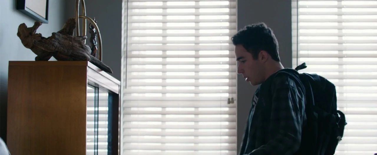 Camp Hideout (2023) movie still: medium — A modern room features a wooden cabinet topped with wooden sculptures against a windowed wall. A young man…