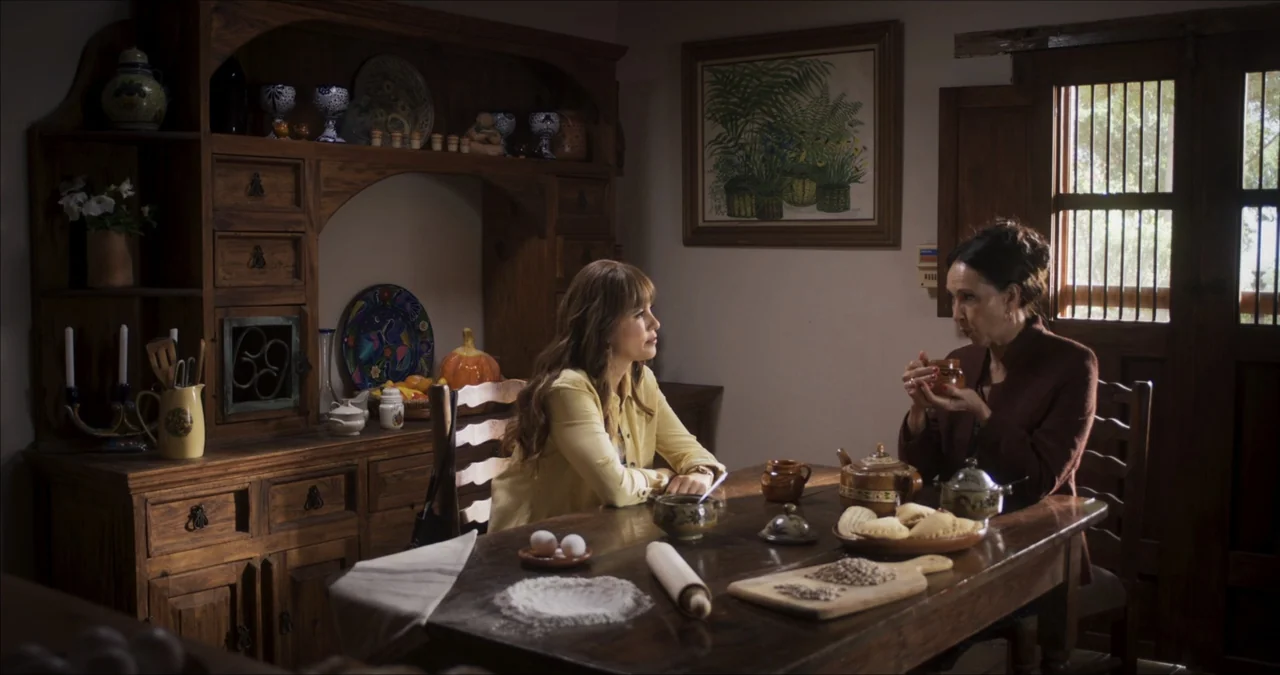 Bendita Suegra (2023) movie still: medium — A rustic dining room features a large heavy wooden table and a matching hutch. Two women sit opposite…