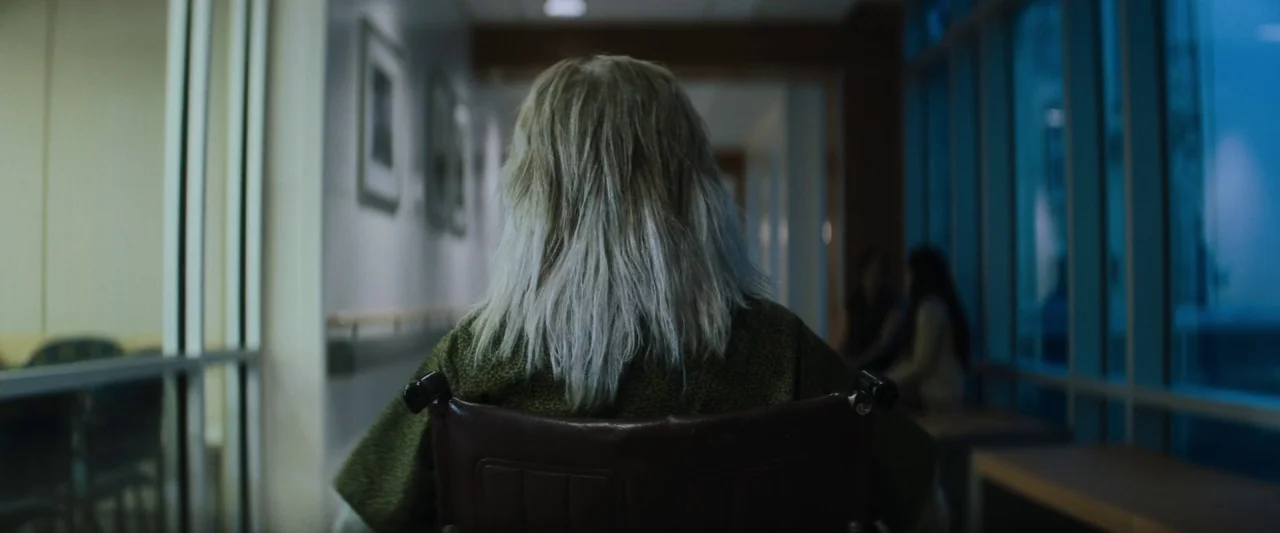 The Grudge (2020) movie still: medium — A long hospital corridor features a glass wall on the right and framed artwork. An elderly woman sits in a…