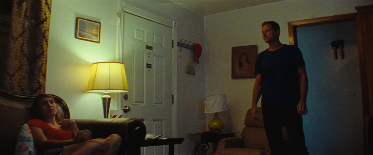 Red Rocket (2021) movie still: wide — A dimly lit room contains two lamps and framed pictures on plain walls. A woman sits on a couch while a man…