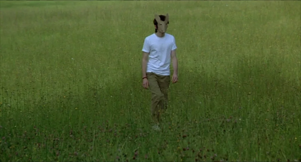On War (2008) movie still: fullbody — A person stands in a large field filled with long green grass and wildflowers. The individual wears a…