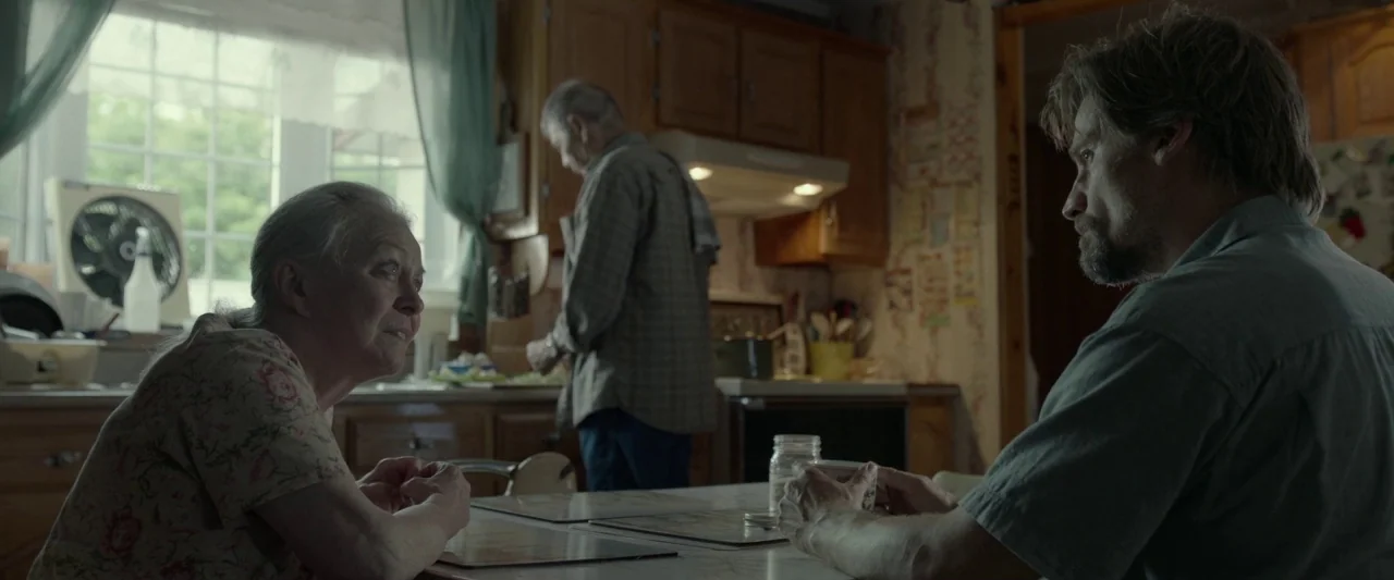 Small Crimes (2017) movie still: medium — A modest dining area features wood cabinets and floral wallpaper. An elderly woman sits across from a…