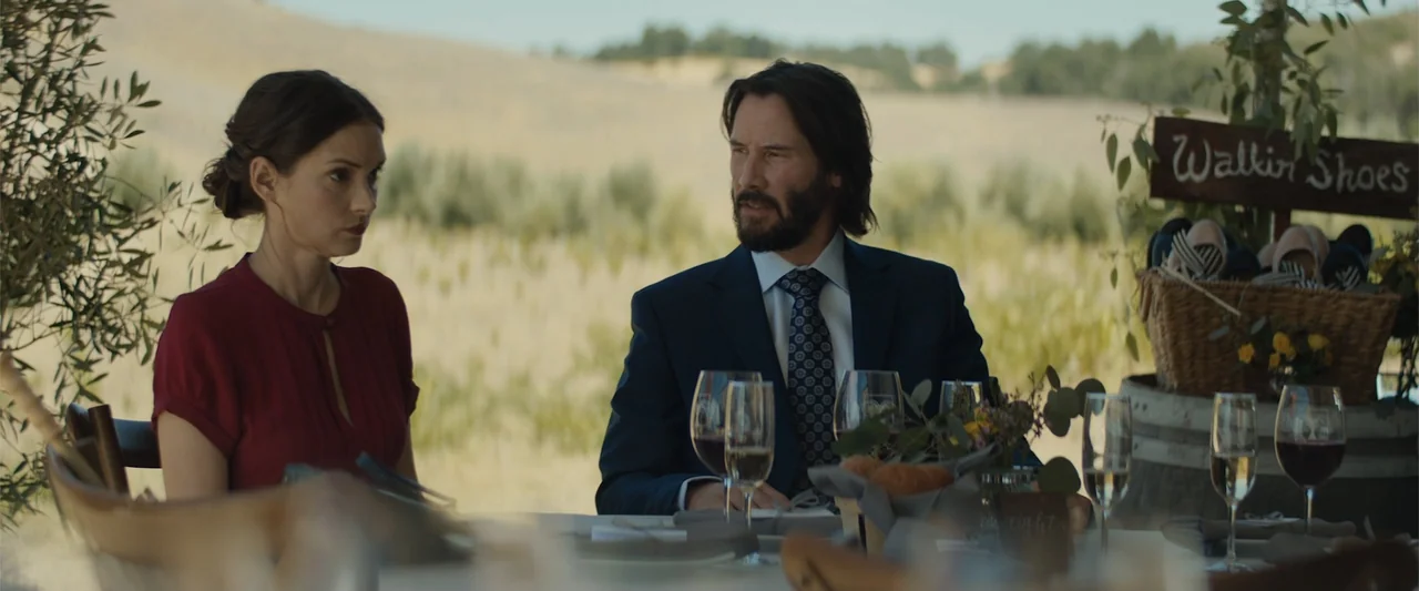 Destination Wedding (2018) movie still: medium — A table is set outdoors in a grassy field with rolling hills in the background. A man in a blue…