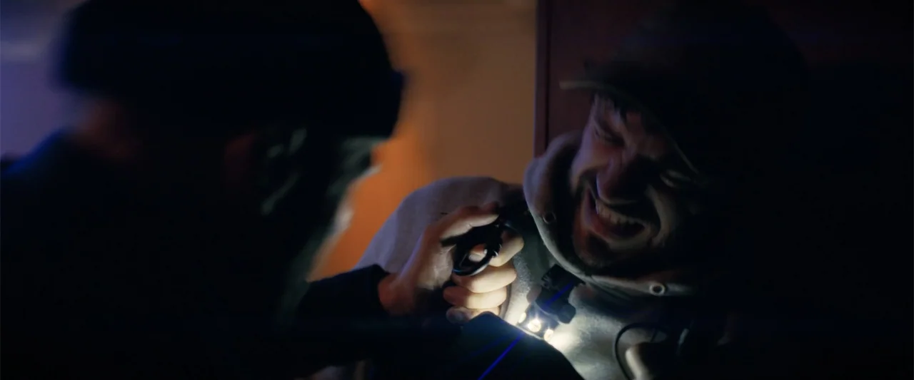 A Working Man (2025) movie still: closeup — A dimly lit indoor space with blurred background walls and shadows. A man in a hoodie is restrained…