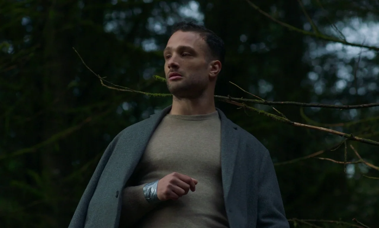 It Is In Us All (2022) movie still: medium — A man stands within a dense, mossy woodland area featuring dark, gnarled tree branches. He gazes…