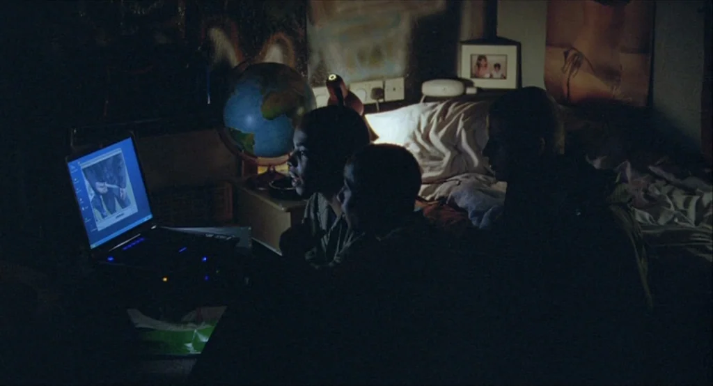 Looking for Eric (2009) movie still: medium — A dimly lit room at night features three young men gathered around a laptop. The room contains a bed,…