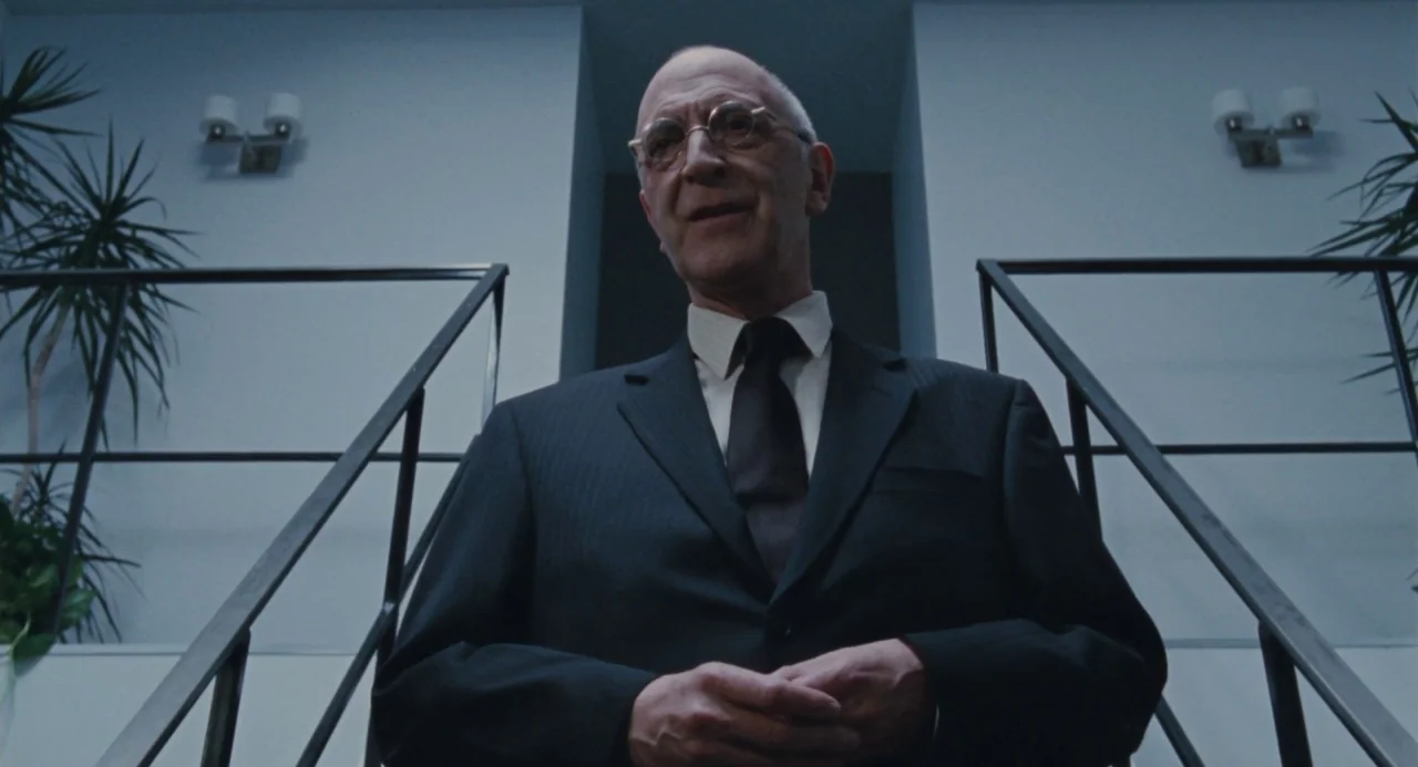 Martyrs (2008) movie still: medium — A man stands atop a staircase in a modern, starkly-lit interior architectural setting. The subject waits while…