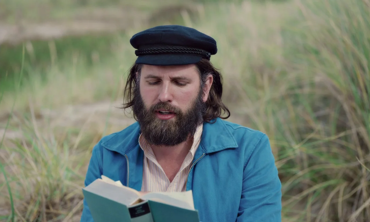 Bloodsuckers (2021) movie still: medium — An outdoor area with tall beach grass and sandy ground under overcast skies. A man with a thick beard…