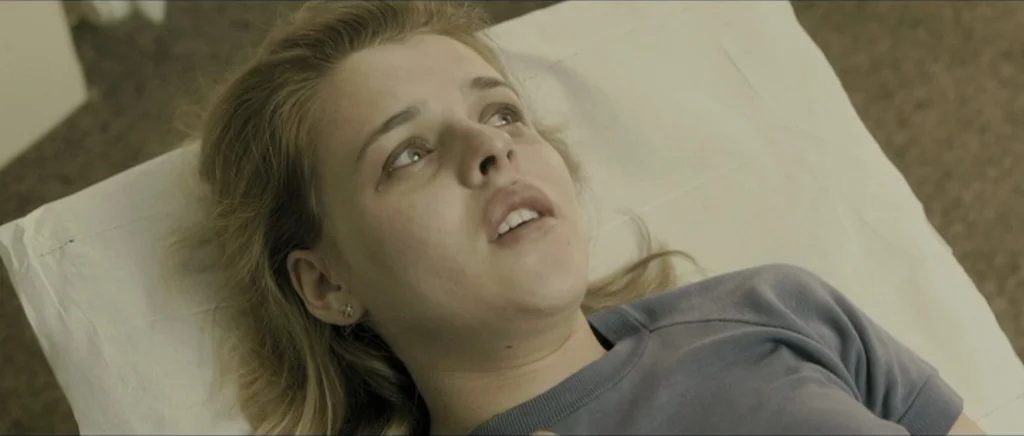 Lasting (Nieulotne) (2013) movie still: closeup — A woman lies on a white pillow atop a clinical surface. She looks upward with a neutral expression…
