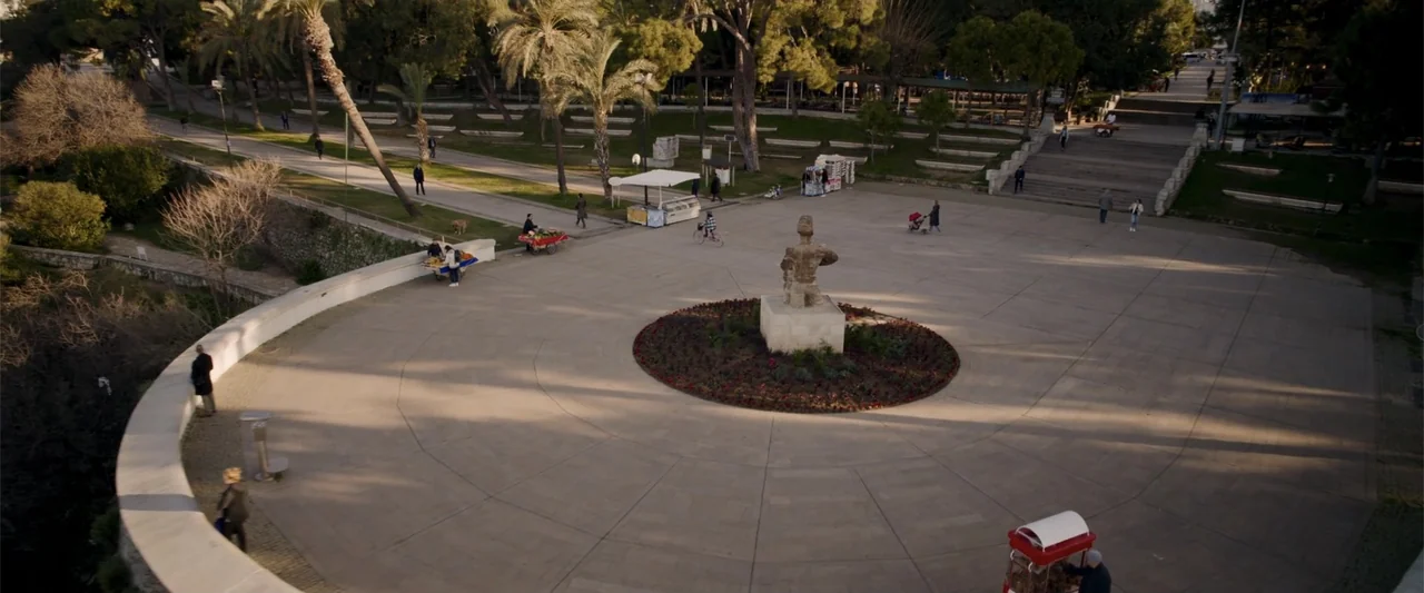 Operation Fortune: Ruse de Guerre (2023) movie still: wide — A spacious stone-paved urban plaza features a central statue and surrounding trees at…