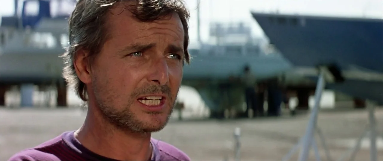 The Parallax View (1974) movie still: closeup — A sunny outdoor boatyard contains multiple boats sitting on metal stands in the background. A man…