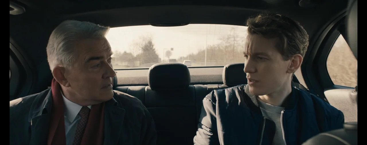 The Wrong Tenant (2022) movie still: medium — The interior of a moving car backseat with two men sitting side by side. An older man in a suit speaks…