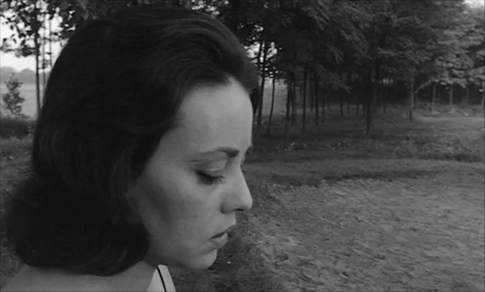 La Notte (1961) movie still: closeup — A woman in a park with rows of trees and a sandy patch. The woman stands facing right with her eyes closed.