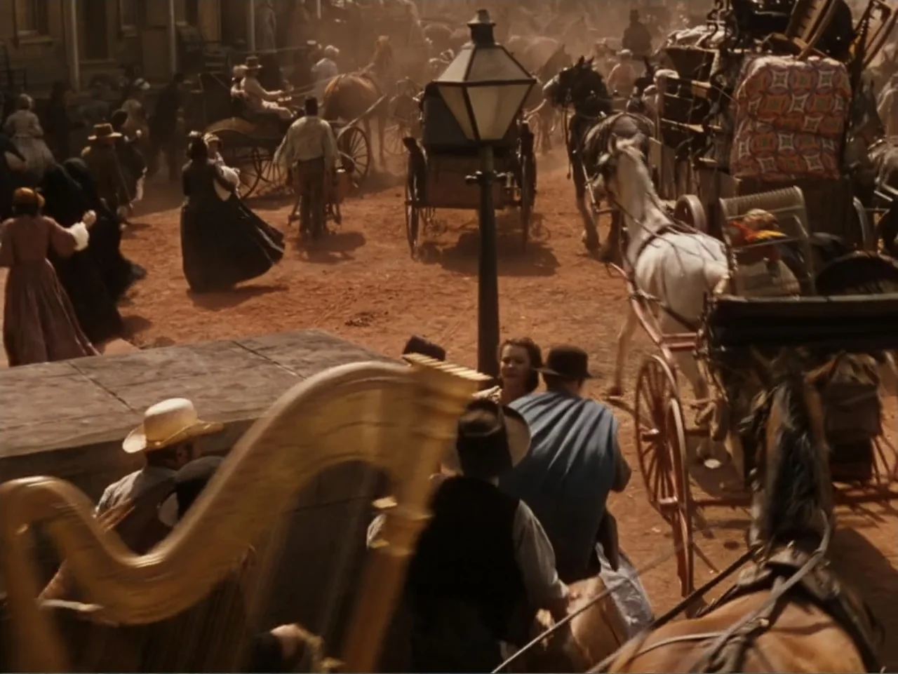 Gone with the Wind (1939) movie still: wide — A crowded 19th-century town square features dirt roads and many horse-drawn carriages. A man and a…