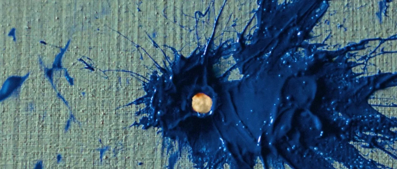 Let The Corpses Tan (2017) movie still: closeup — Thick navy blue paint is splattered across a coarse beige canvas. A small circular hole pierces…