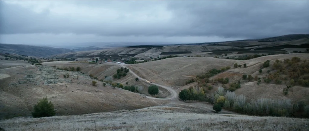 Once Upon a Time in Anatolia (2011) movie still: establishing — A vast, rolling landscape of dry hills under a thick layer of grey clouds. A narrow,…
