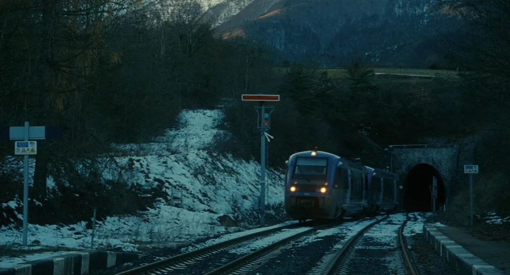 Things to Come (2016) movie still: wide — A passenger train moves along tracks toward the dark opening of a stone tunnel. The surrounding landscape…