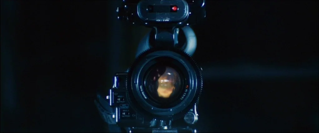 Kick Ass (2010) movie still: closeup — A professional video camera sits in a pitch-black environment facing the camera directly. The glass lens…