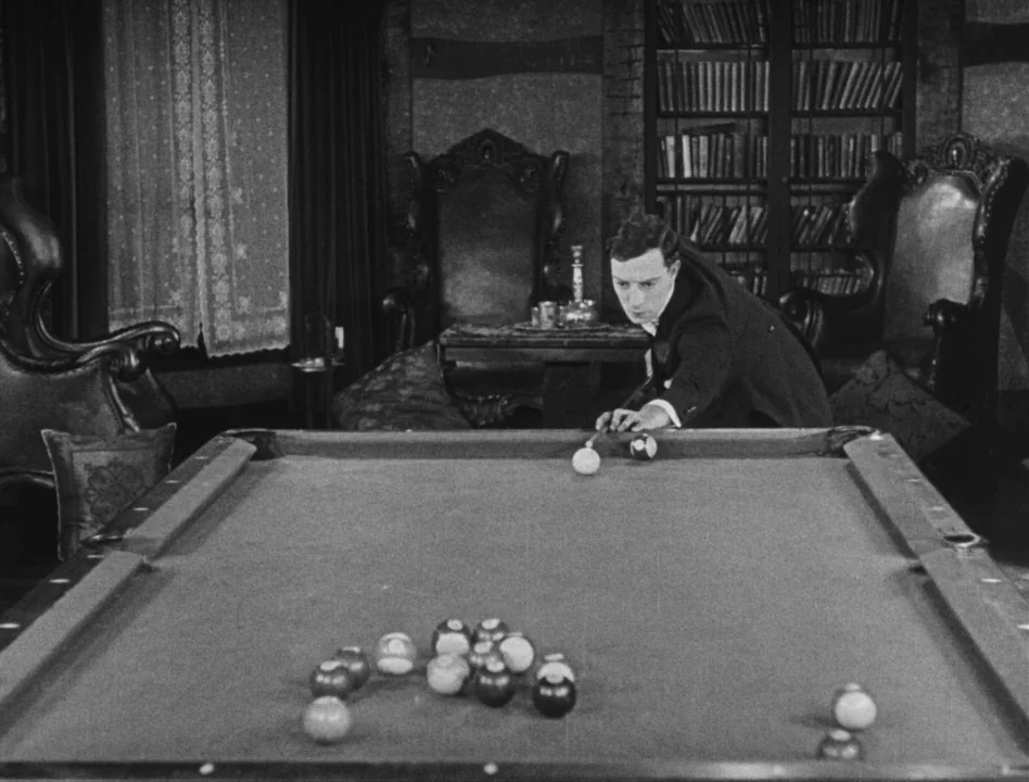 Sherlock Jr. (1924) movie still: medium — A vintage billiards room features a heavy pool table, bookshelves, and dark leather armchairs. A man in a…