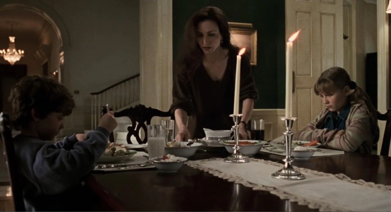 Jumanji (1995) movie still: medium — A dark wood dining table set for three people in an upscale home interior. A woman serves food to a young boy…