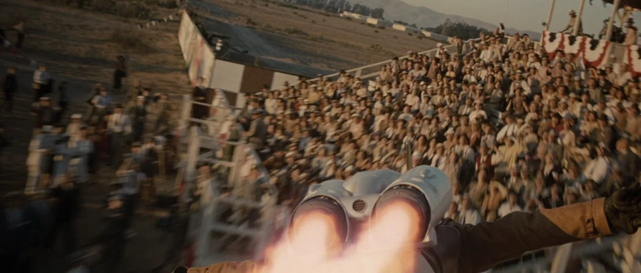 The Rocketeer (1991) movie still: wide — A dense crowd of people sits in wooden bleachers decorated with festive red and white bunting. A pilot…