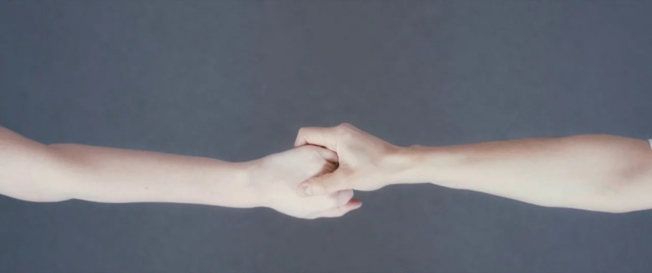 Ginger & Rosa (2012) movie still: closeup — A featureless, neutral grey surface serves as the backdrop for two hands. Two human arms reach from…