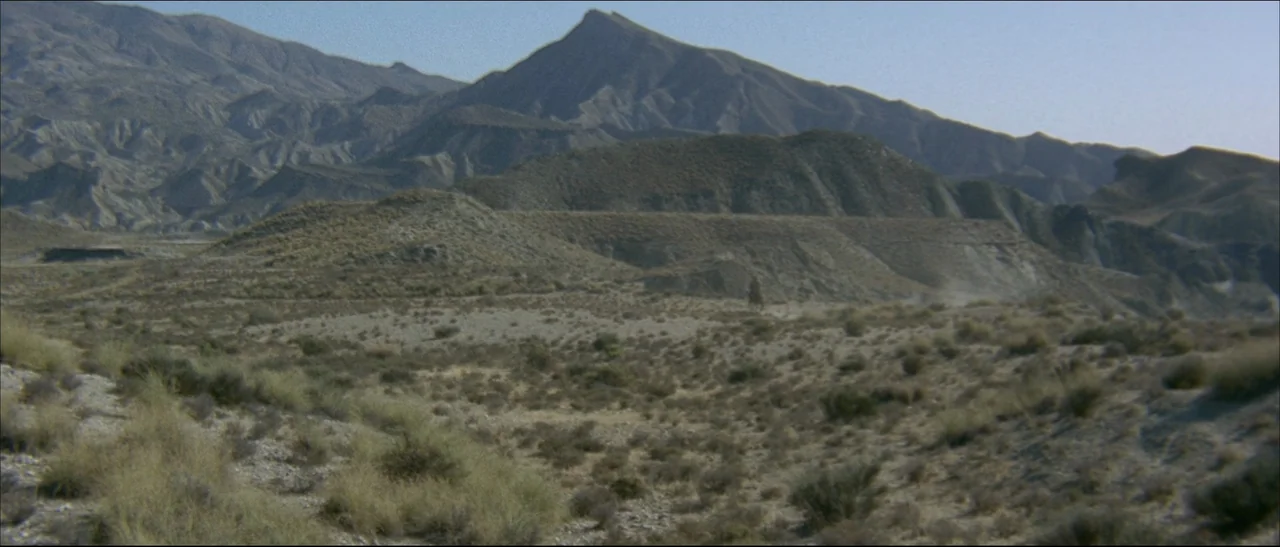 The Mercenary (1968) movie still: establishing — An arid, rocky desert landscape stretches toward rugged mountain peaks in the distance. Sparse dry…