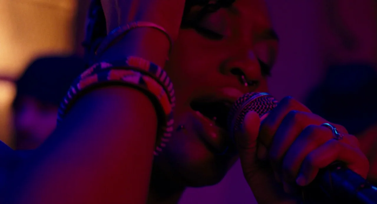 Pariah (2011) movie still: closeup — A dark indoor music venue filled with neon purple and red stage lights. A woman performs at a microphone while…