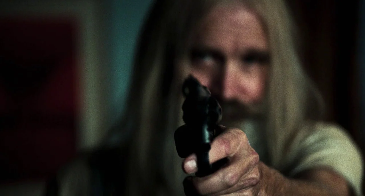3 From Hell (2019) movie still: closeup — A dimly lit room with muted blue tones in the shadows. A man with long hair points a black handgun…