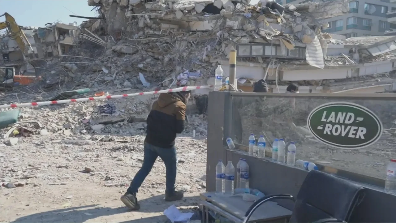 Death Without Mercy (2024) movie still: wide — A destroyed building lies in ruins with piles of concrete and twisted metal debris. A man in a brown…