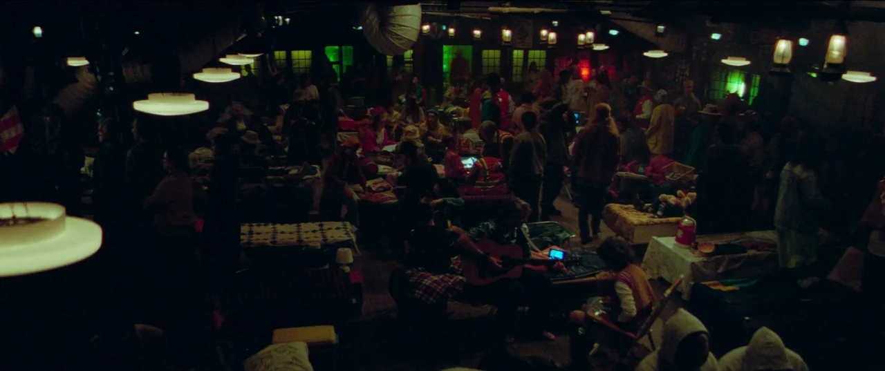 White Noise (2022) movie still: establishing — A dark indoor space filled with numerous cots and people sitting on the floor. Dim pendant lights and…
