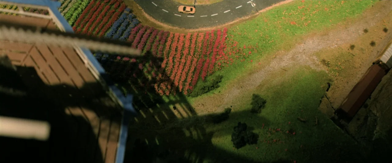 Holland (2025) movie still: establishing — A high-angle view looks down at a miniature model landscape featuring rows of colorful flowers. A small…