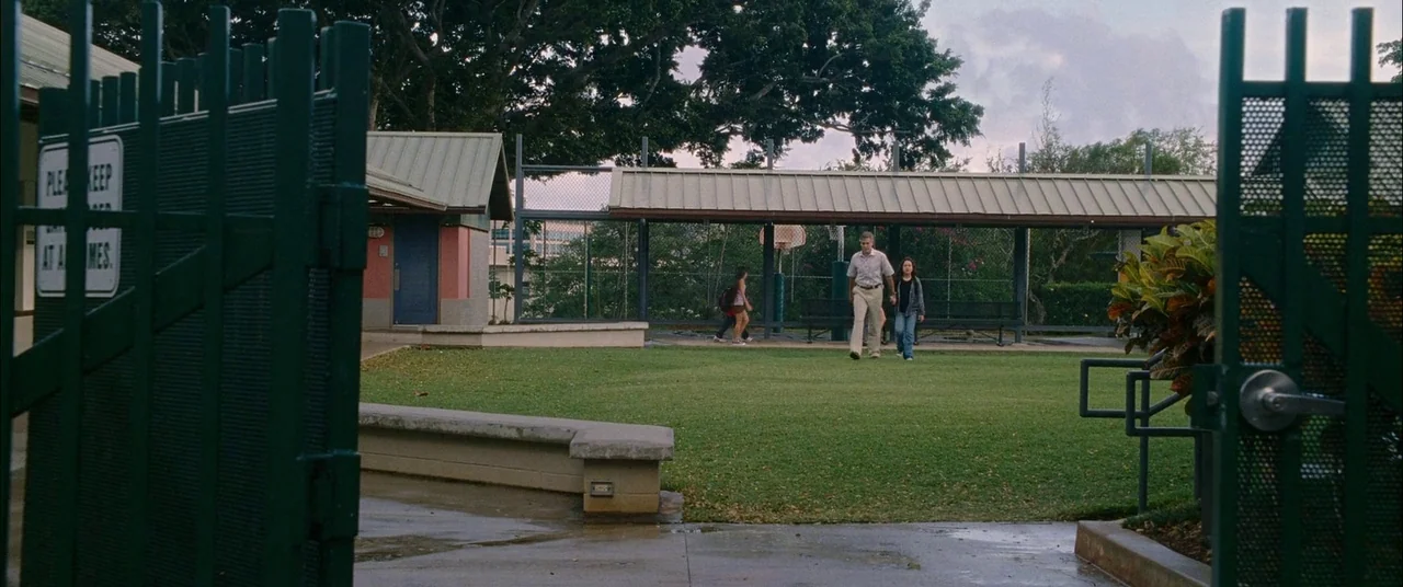 The Descendants (2011) movie still: wide — A school courtyard features a manicured lawn, a covered walkway, and green metal gates. An older man…