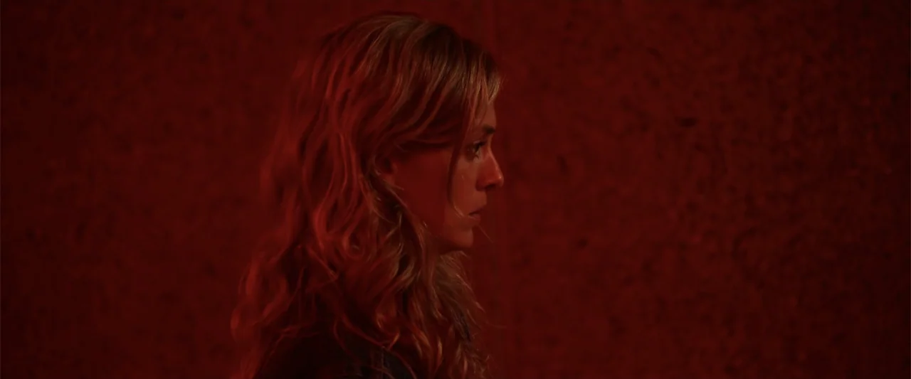 Init!ation (2021) movie still: closeup — A dimly lit room features a textured dark red wall as the backdrop. A woman with wavy blonde hair stands in…