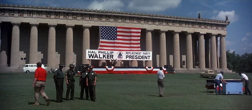 Nashville (1975) movie still: wide — A wide shot shows a political rally stage set up before a large columned building. Several men stand on a lawn…