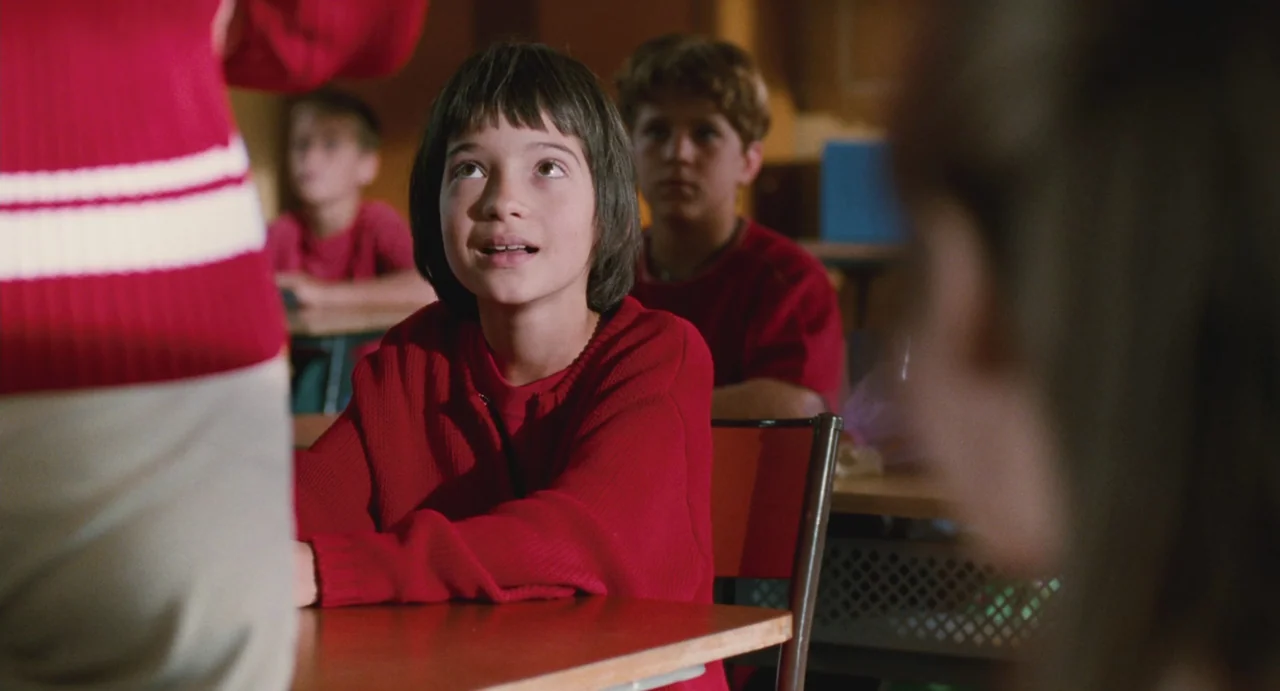 Ma Vie En Rose (1997) movie still: medium — A student sits at a wooden desk in a bright classroom with others behind. A young girl in a red sweater…