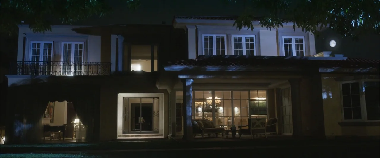 Crazy for Change (2020) movie still: wide — A large two-story suburban mansion at night featuring a lit porch and visible interior rooms. Exterior…