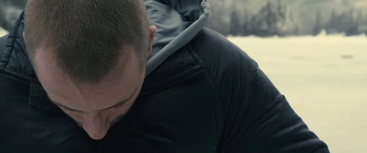Rust And Bone (2012) movie still: closeup — A man stands outdoors on an expansive, white frozen lake covered in light snow. He looks downward toward…