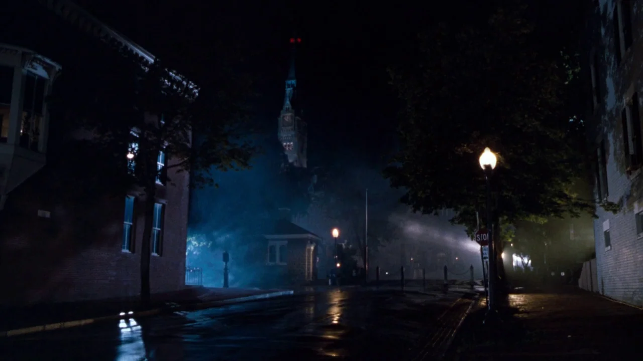 The Exorcist 3 (1990) movie still: establishing — A quiet town square is filled with low-hanging fog at night. A historic clock tower silhouette…