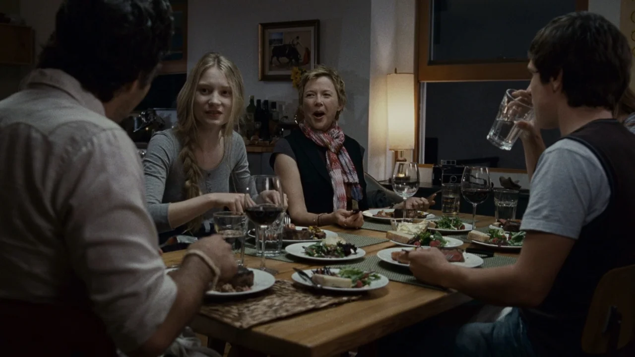 The Kids Are All Right (2010) movie still: medium — A group of people sits at a wooden dining table in an interior room at night. They are eating a…